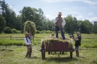 Two farmers pack a cart with hay while another person films