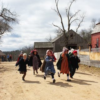A group of kids wearing 1830s style clothes run down a dirt road