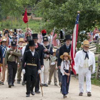A group of people dressed in 1830s attire parade in the countryside, let by a girl carrying a large American flag