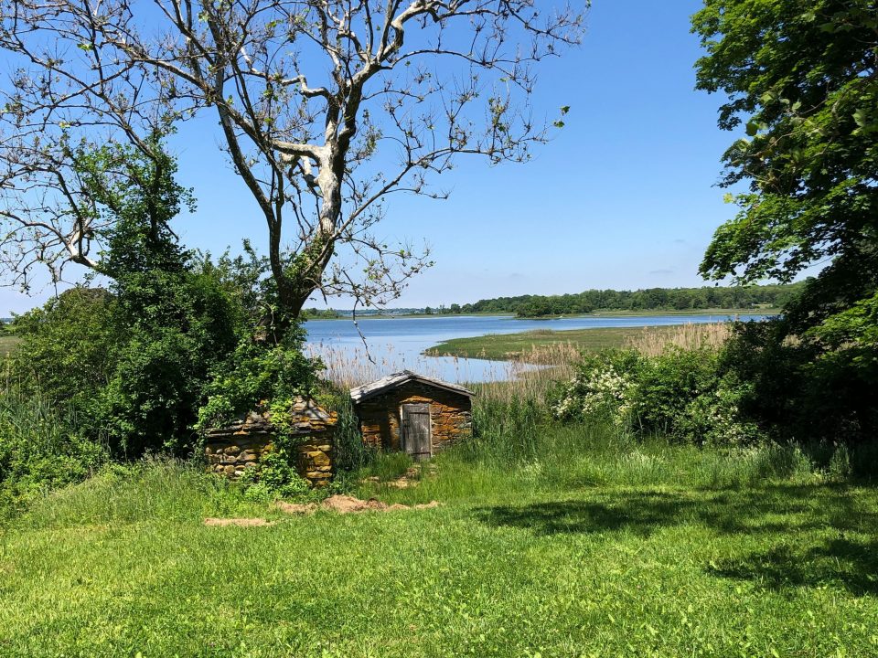 An outside photos showing two small stone structures, grass, and the bay at a distance