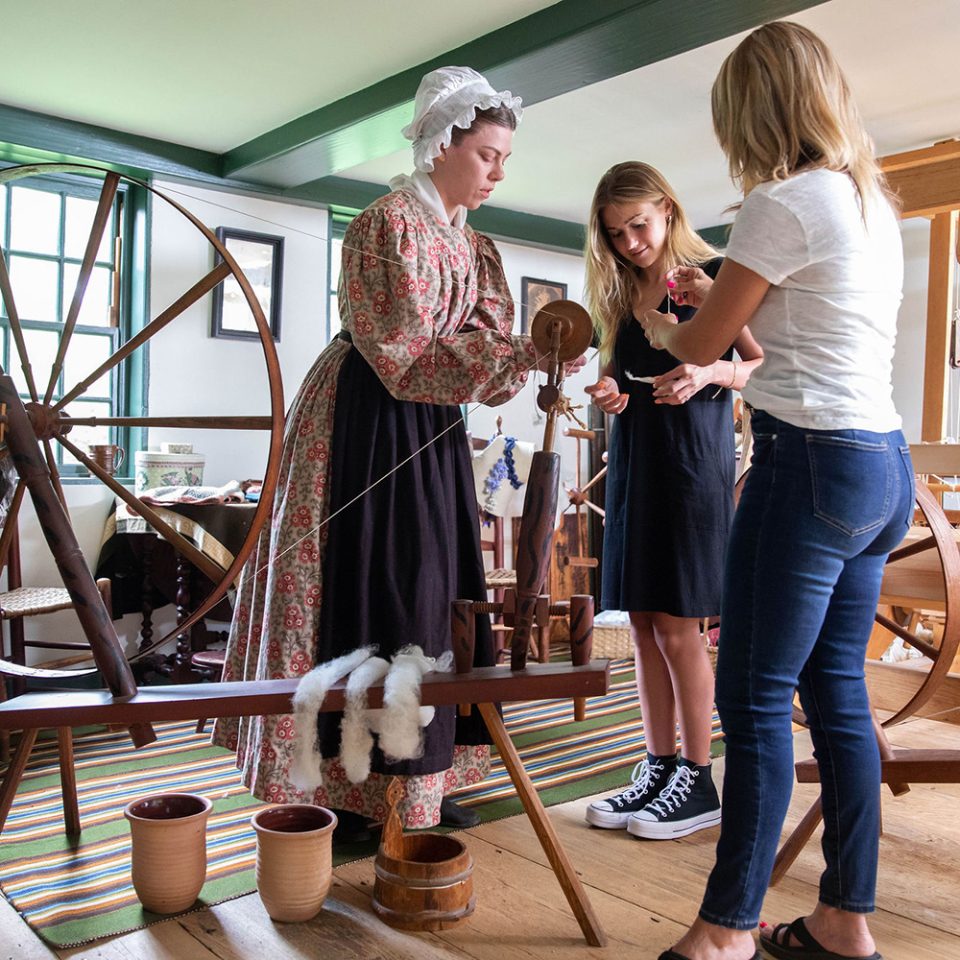 Two visitors chat with a costumed historian and try their hands at spinning yarn