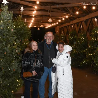 Three people stand for a photo on a covered bridge filled with lit up Christmas trees